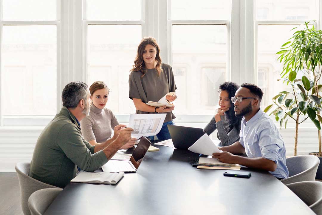 Five colleagues collaborate at a conference table with laptops and documents in a bright office