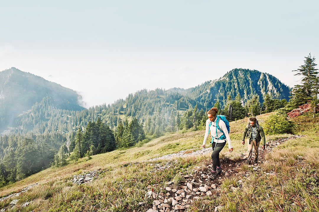 A couple hiking through a field with backpacks on and mountains in the background